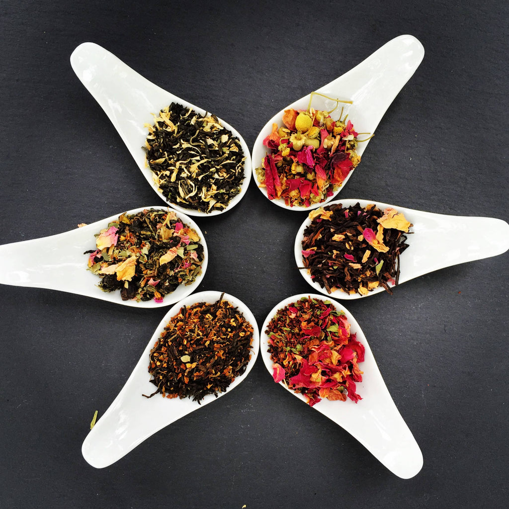 Assorted TOCHA organic loose-leaf tea blends with floral and herbal ingredients displayed in white spoons on slate background